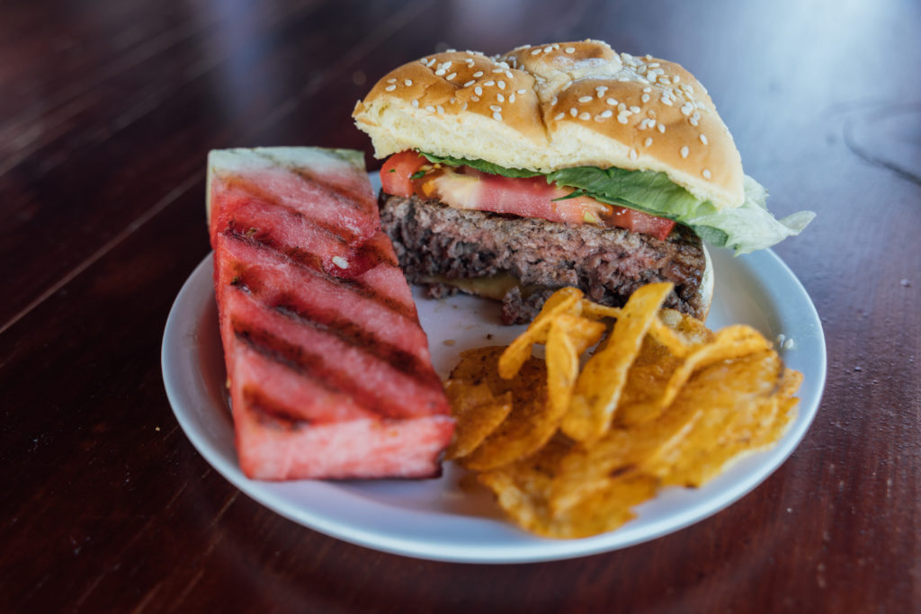 A hamburger plated where you can see the doneness of the burger with the grilled watermelon and kettle chips.