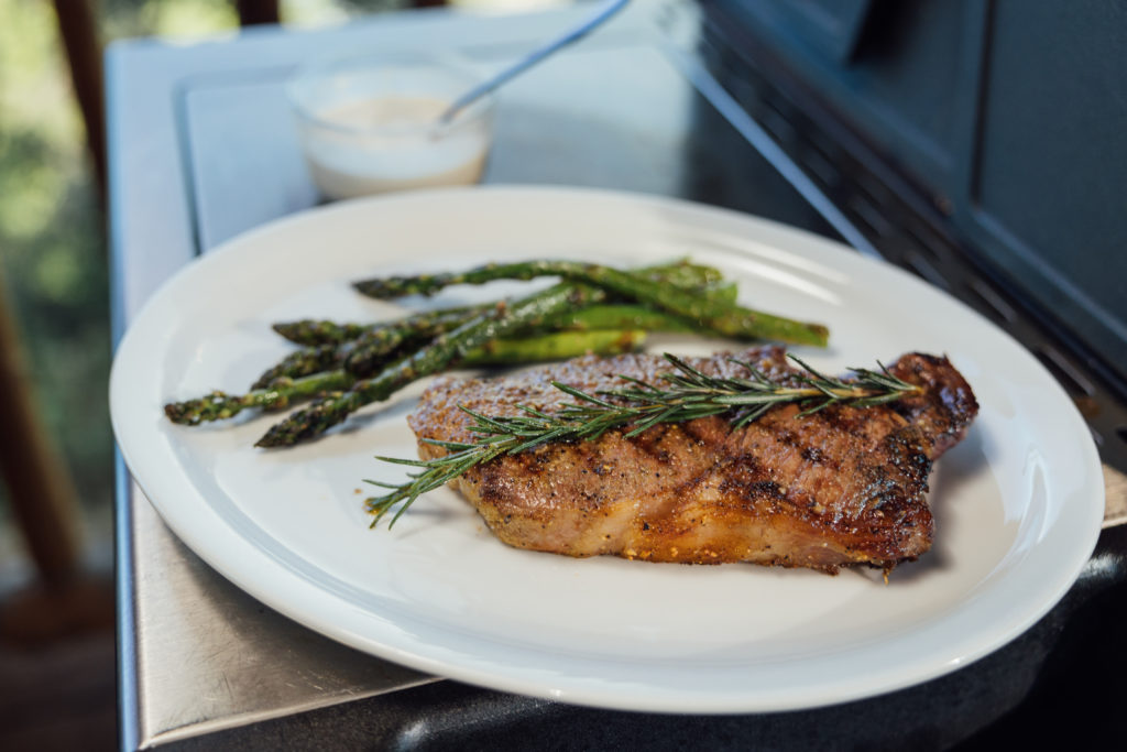 New York Strip Steak is grilled with a piece of rosemary over the top. Plated next to it is some grilled asparagus and you can see the creamed horseradish in a serving dish behind the plate.